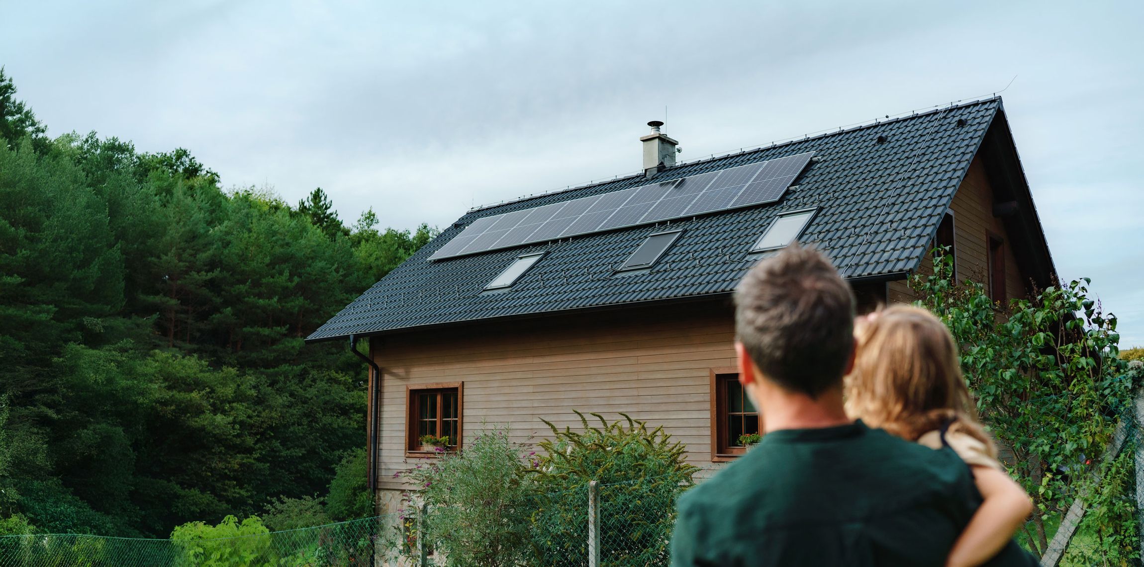 Zwei Personen von hinten blicken auf ein Haus mit Solarpaneelen auf dem Dach vor bewaldetem Hintergrund